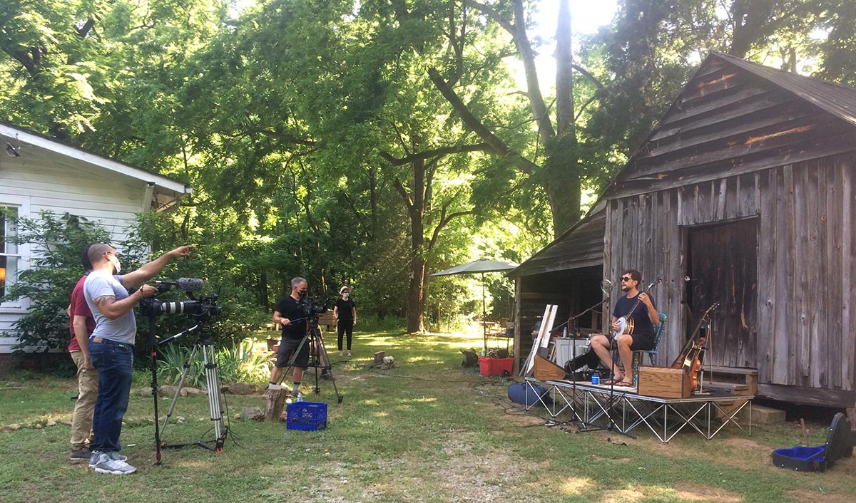Camera operators discuss filming while a musician sits with a banjo on on outdoor stage