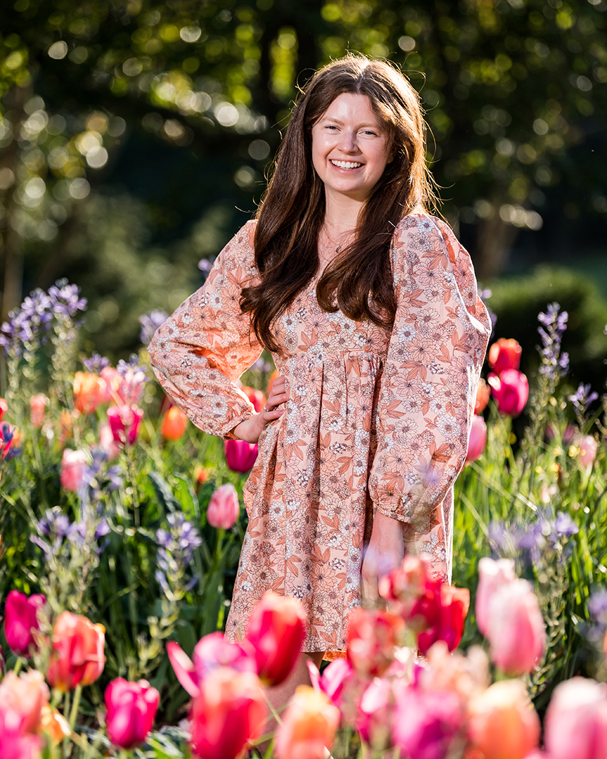 A woman in a pink dress, with long brown hair, smiling as she stands among pink and orange tulips and other colorful flowers.