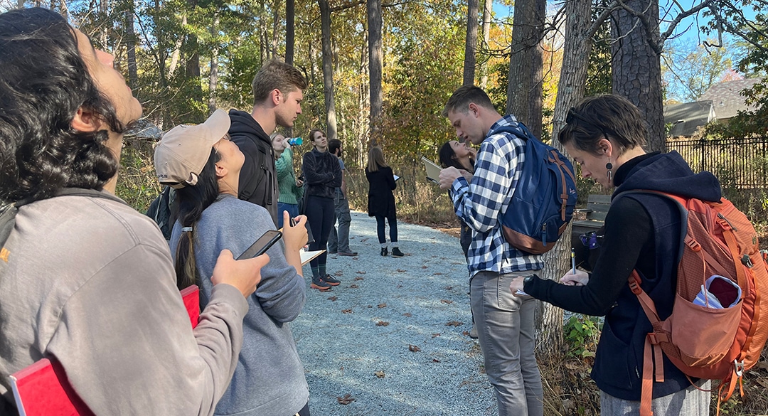 8 college-age people stand on a path among trees, some looking up and some writing in notebooks in their hands.