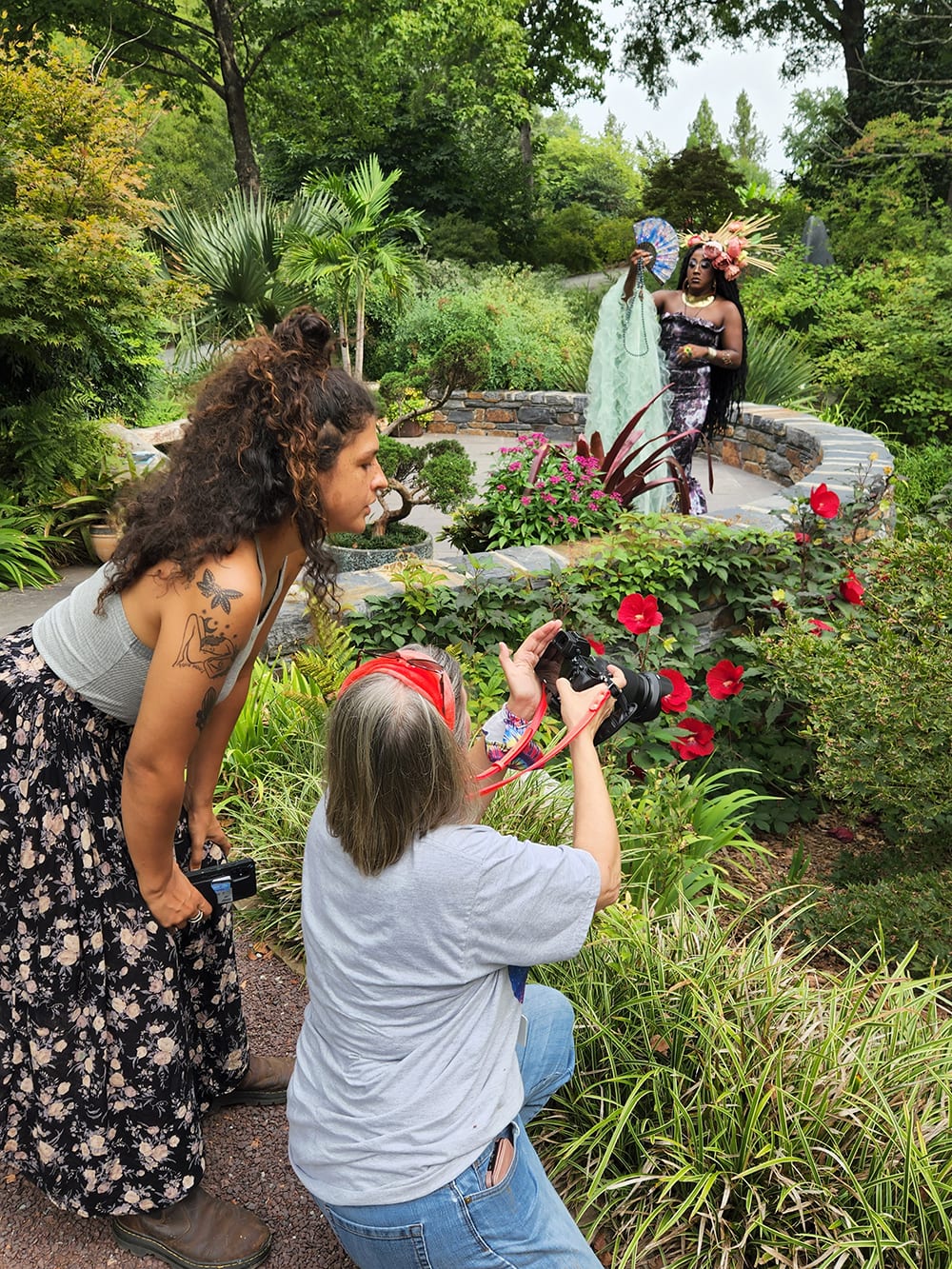 Two women confer while one photographs a person in a long purple dress and holding a fan and a pale green scarf, all standing in a verdant garden with red flowers and lots of varied greenery.