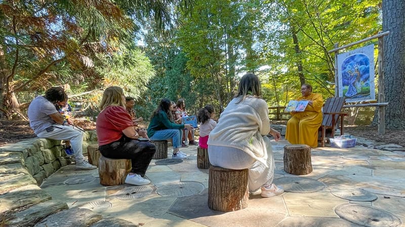 A group of people of various ages sit on tree stumps outdoors while another person sitting in a chair holds a book while reading to them.