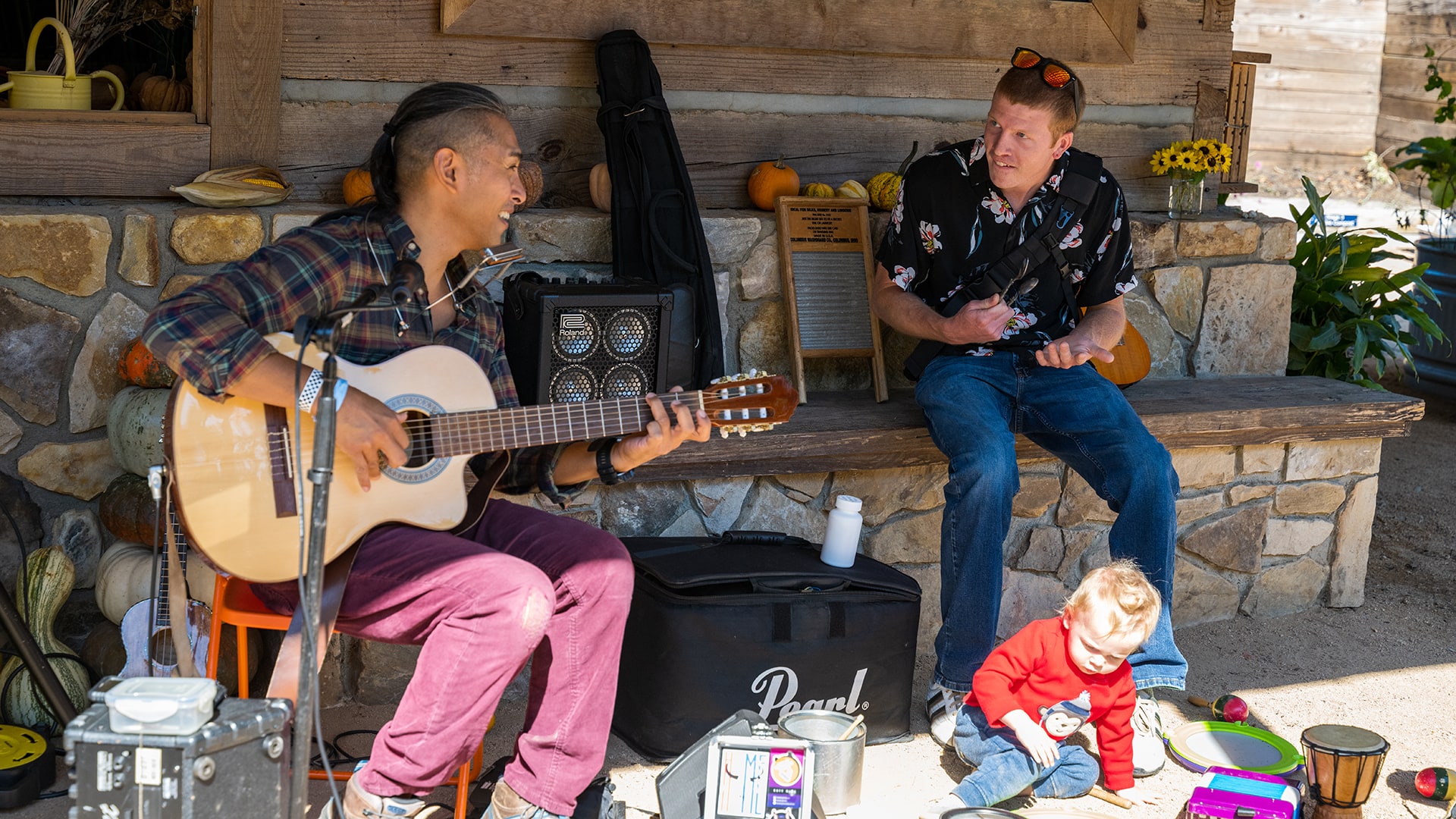A man with a guitar laughs as another man seated nearby gestures to him and a little boy sits on the ground below.