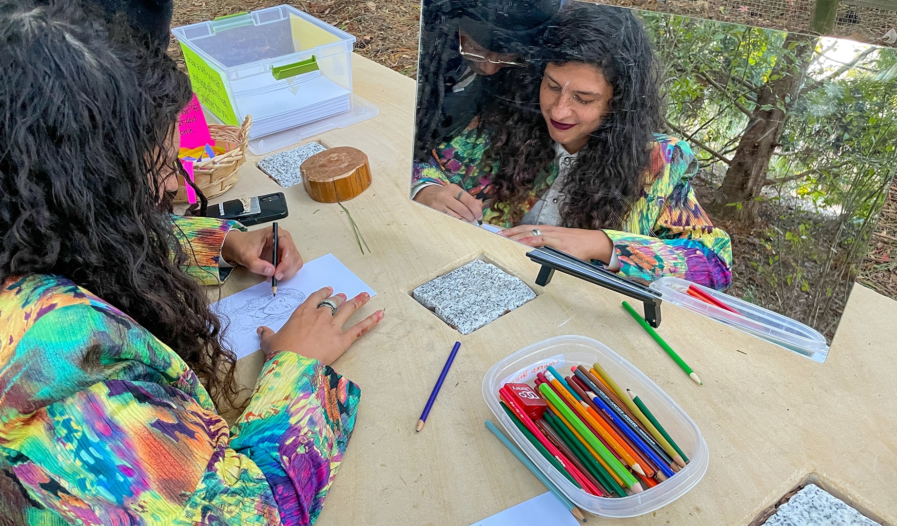 A young woman in a colorful jacket draws a self-portrait with a pencil, surrounded by art supplies and a mirror.